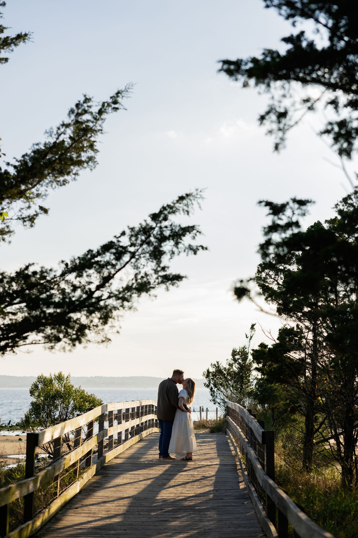 A couple shares a romantic moment on a wooden boardwalk overlooking the ocean during sunset.