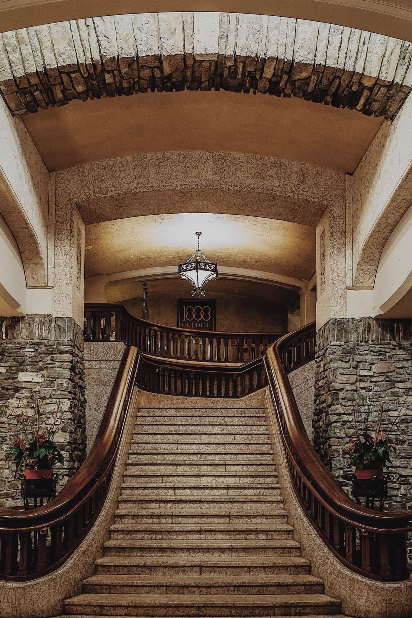 Elegant grand staircase with curved wooden railings and stone walls in a vintage mansion featuring an ornate chandelier.