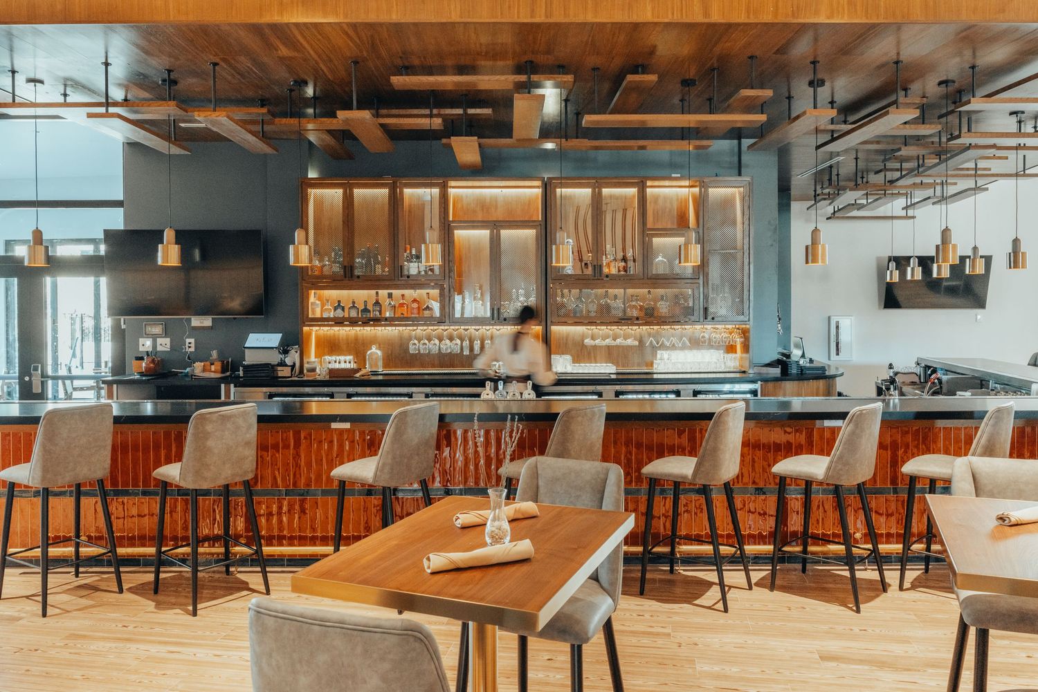 Modern restaurant bar interior with wood tables, tan leather stools, and industrial lighting fixtures in warm tones.