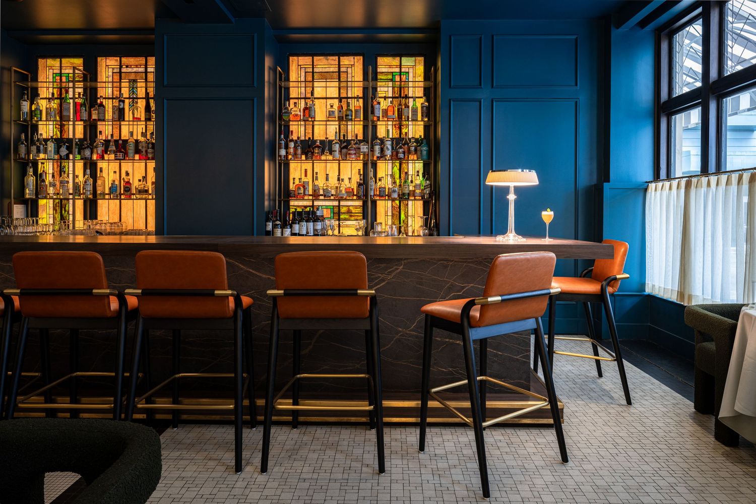 Moody bar interior with leather barstools and backlit liquor shelves glowing against deep blue walls.