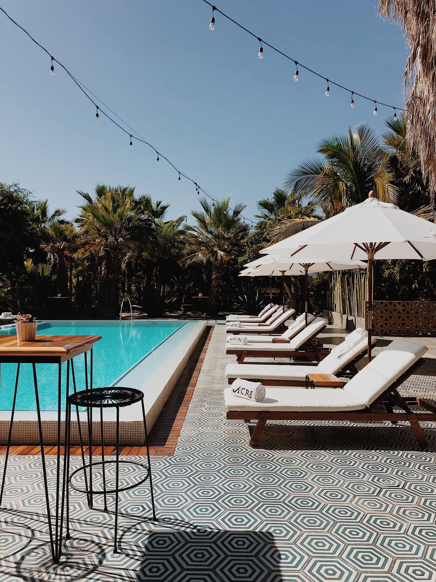Luxurious pool deck with white lounge chairs, patterned tile flooring, string lights, and tropical palm trees at a resort.