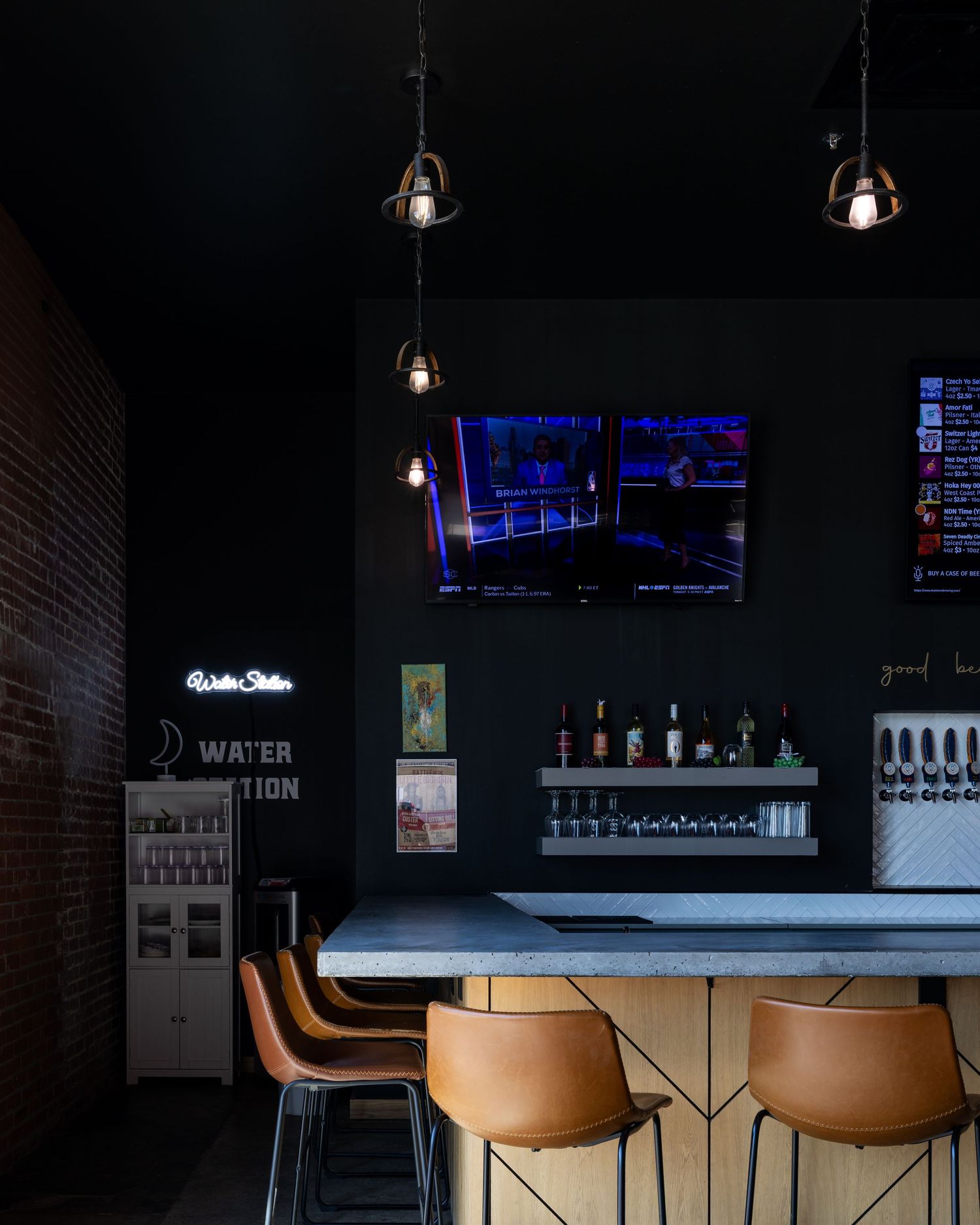 Modern bar interior with exposed brick wall, hanging pendant lights, leather barstools and blue-lit TV displays mounted above.