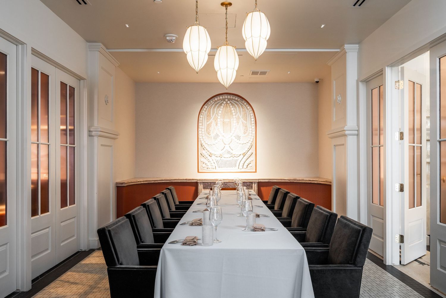 An elegant private dining room with a long white table, black chairs, and hanging pendant lights beneath an arched wall design.