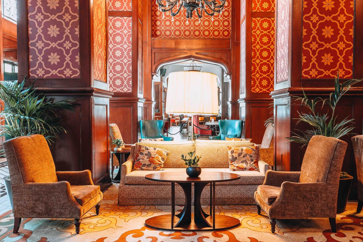 Luxurious hotel lobby lounge with ornate red wallpaper, velvet armchairs, and elegant lighting fixture above couch seating.