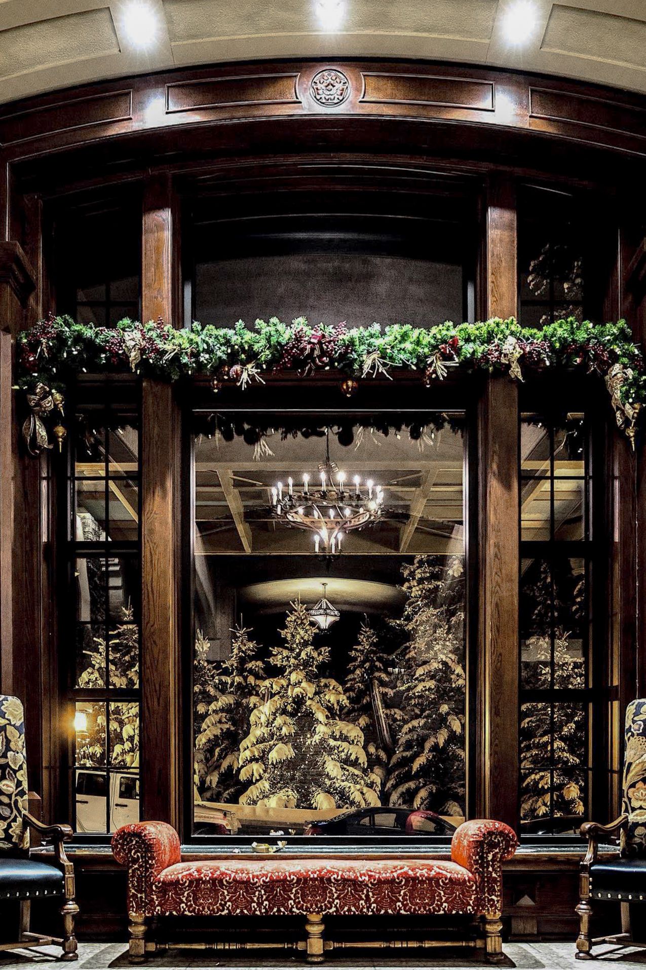 Elegant bay window alcove with vintage leather bench, crystal chandelier, and holiday greenery garland in luxurious interior.
