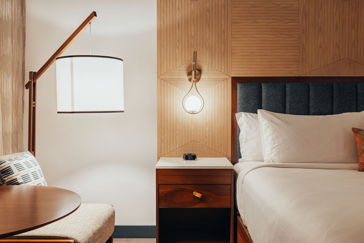 Modern hotel room interior with wooden wall panels, white bedding, and stylish pendant lamp lighting alongside bed.