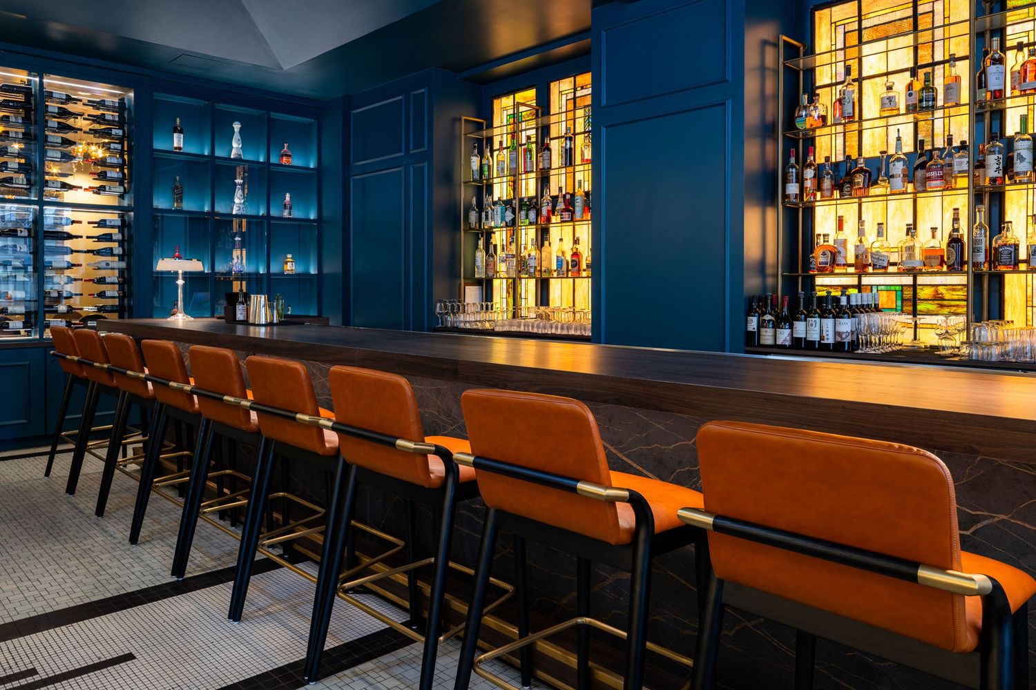 Modern bar interior with illuminated liquor shelves and orange leather barstools along a sleek counter.
