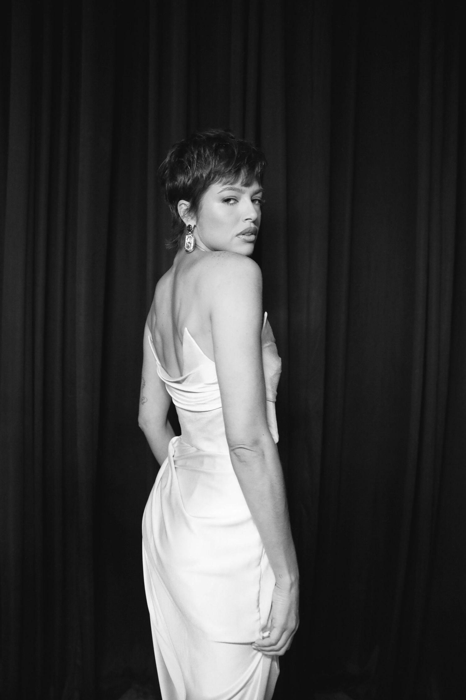 A glamorous black and white photo shows a woman in an elegant white dress posing against a dark curtain background.