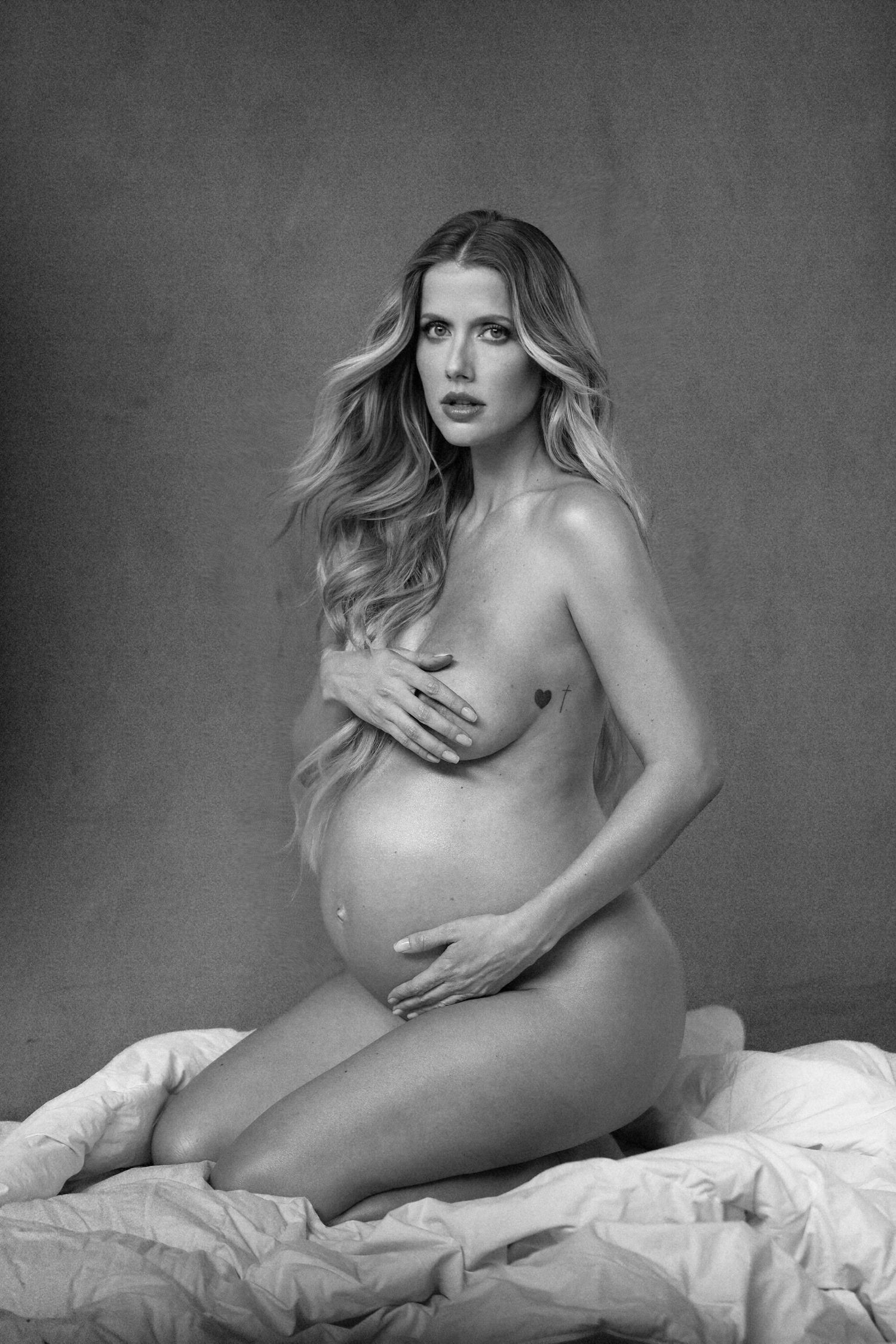 Artistic black and white maternity photo of a pregnant woman posing nude against a simple background.