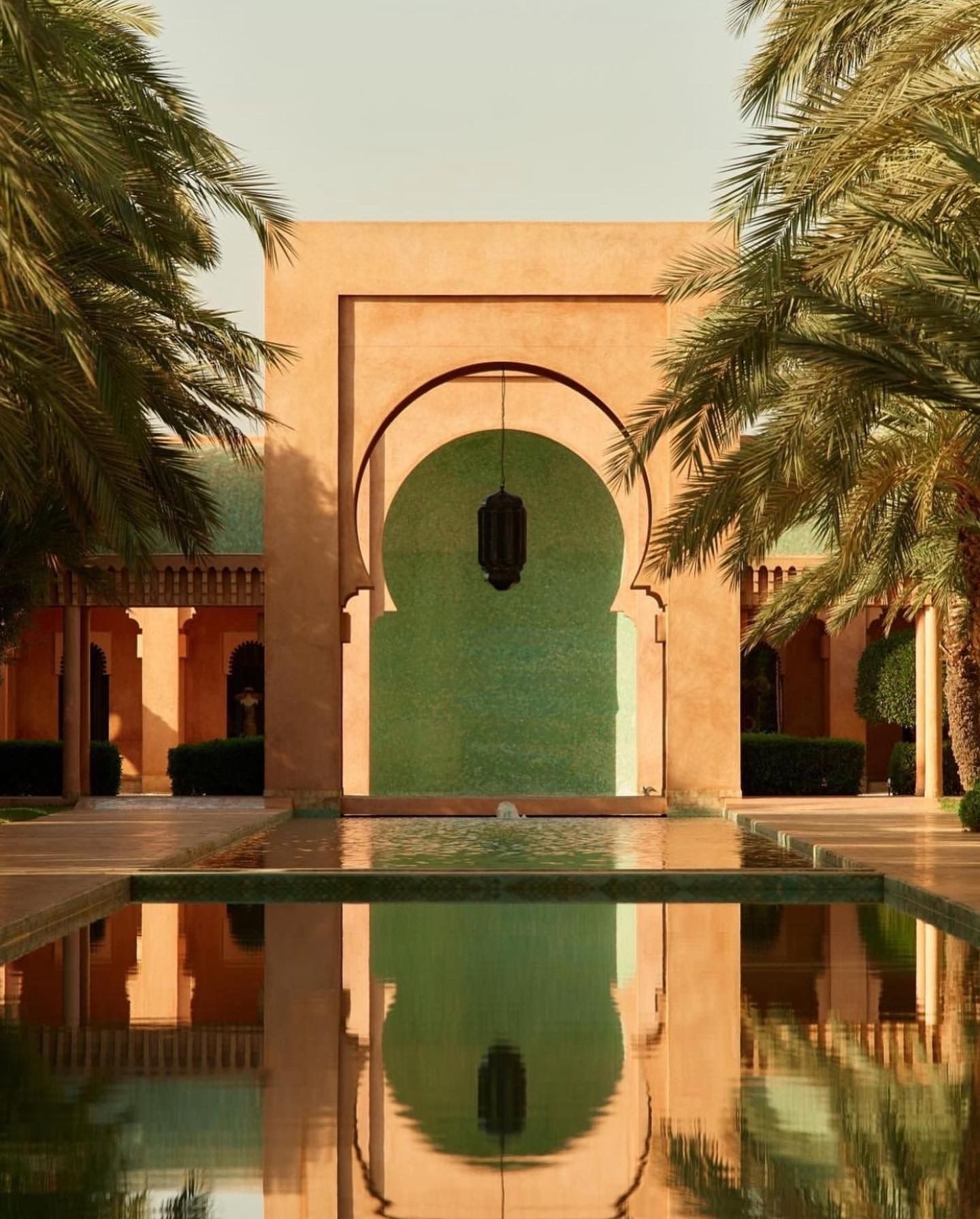 Moroccan-style archway and pool reflecting palm trees and peach-colored architecture in a serene courtyard setting.
