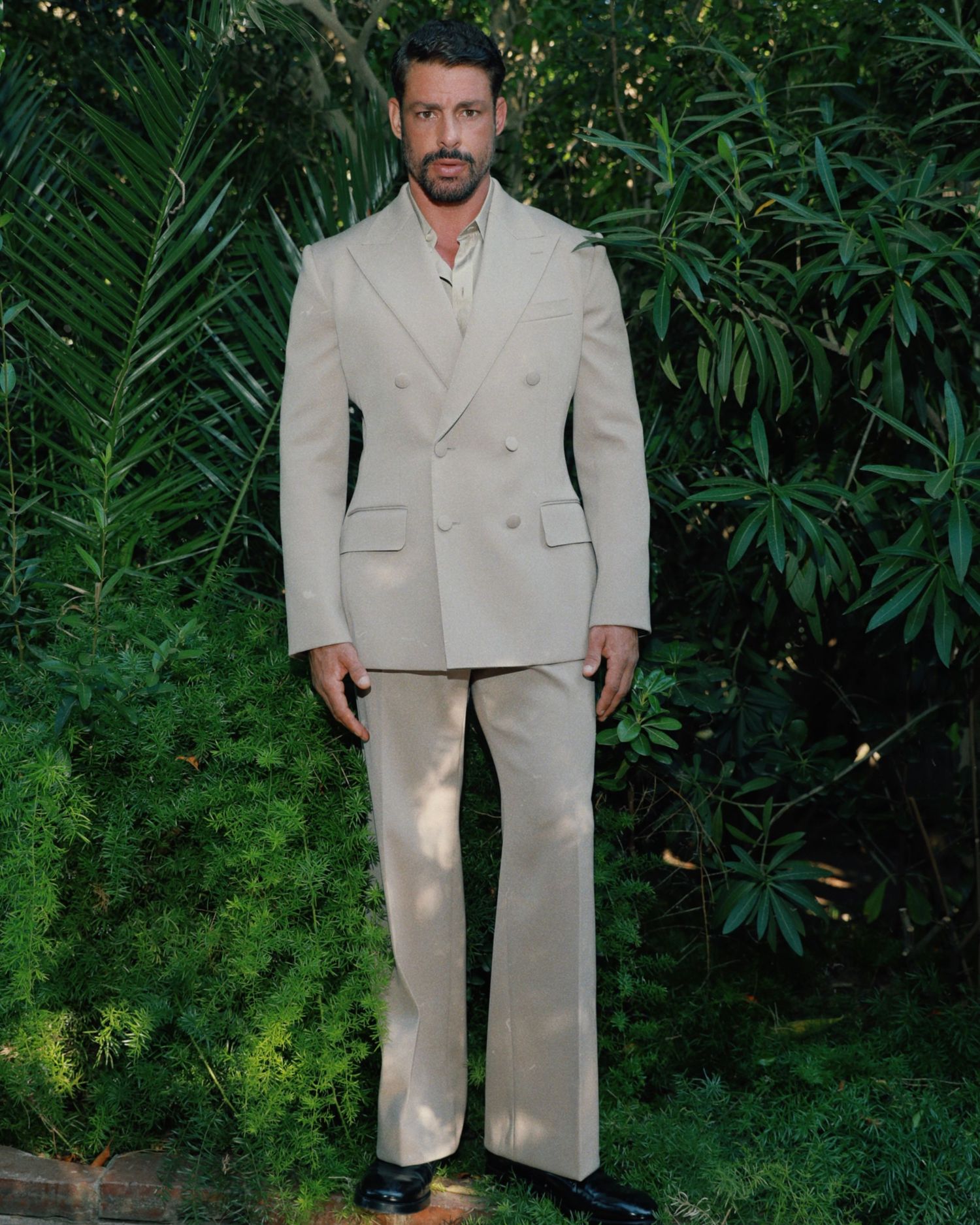 A person wearing a beige double-breasted suit stands in front of lush green foliage in an outdoor setting.