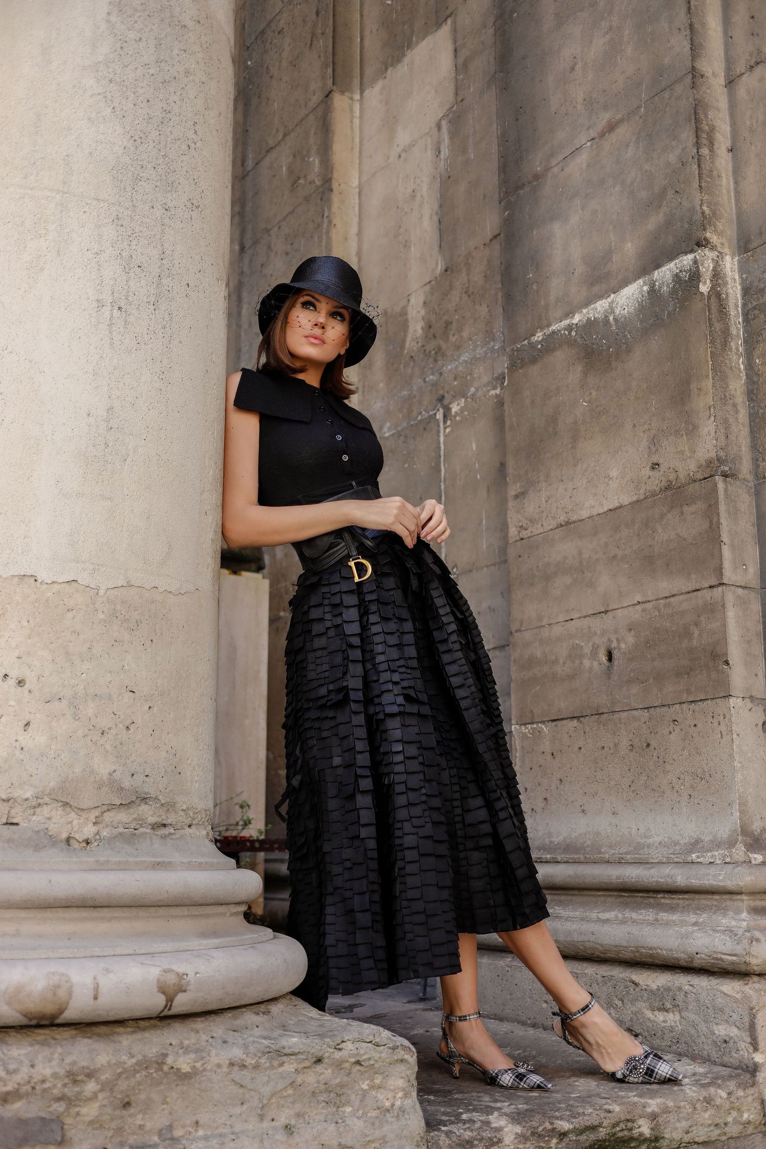 A person in a vintage-inspired black outfit with bowler hat poses elegantly against classical stone columns.