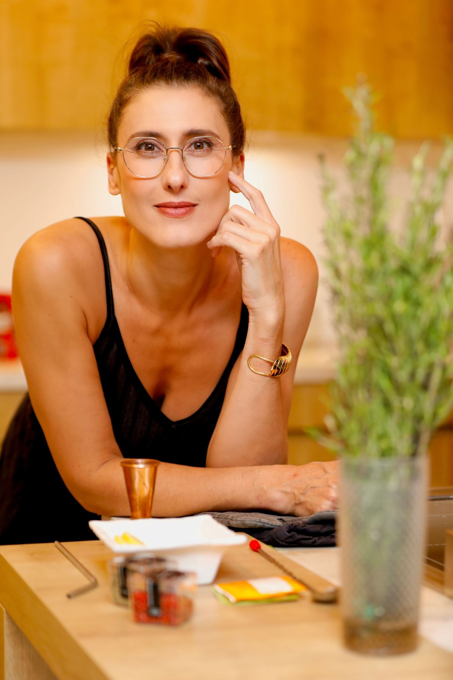 Woman in black tank top and glasses sitting casually at table with indoor plants.