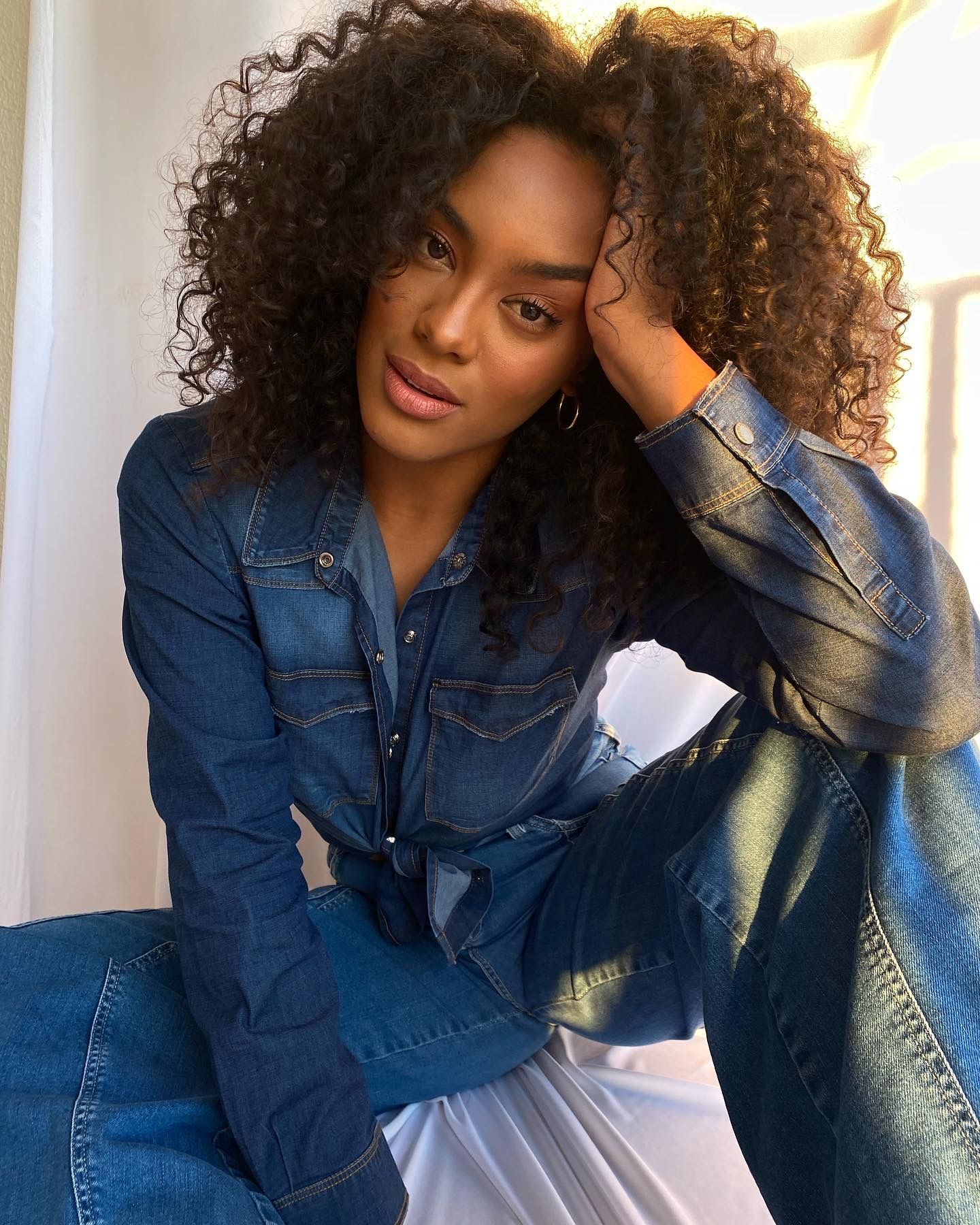 Portrait of woman in denim outfit with natural curly hair against white background.