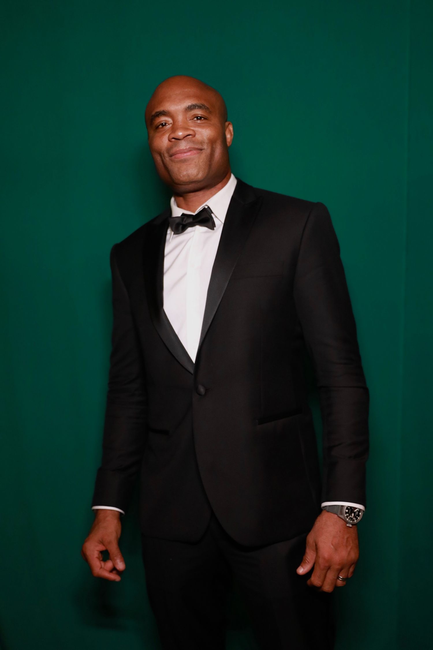 A man in a black tuxedo and bow tie poses against a dark green background.