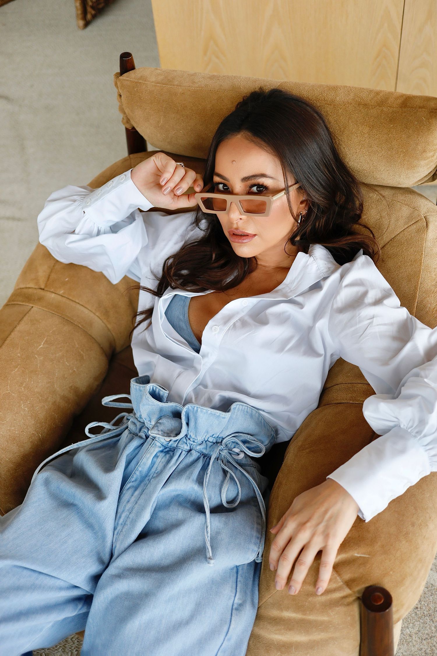 Fashion model lounging on tan couch wearing white button-up shirt and light denim.