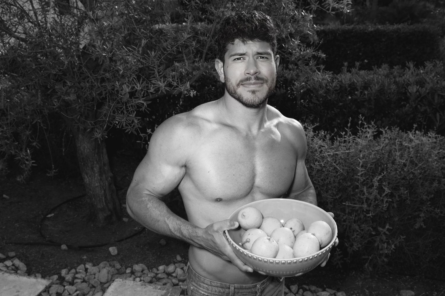 Black and white photo of a shirtless muscular person holding a bowl of eggs outdoors in a garden setting.