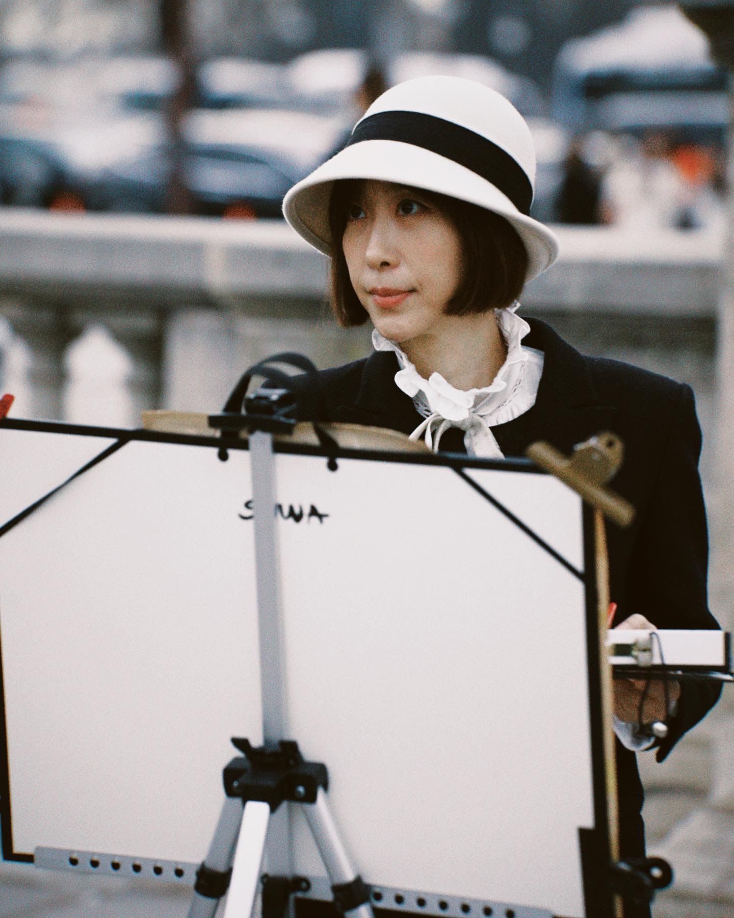 Stylish figure in a black blazer and white fedora hat stands at an easel or display board outdoors in vintage-style photo.