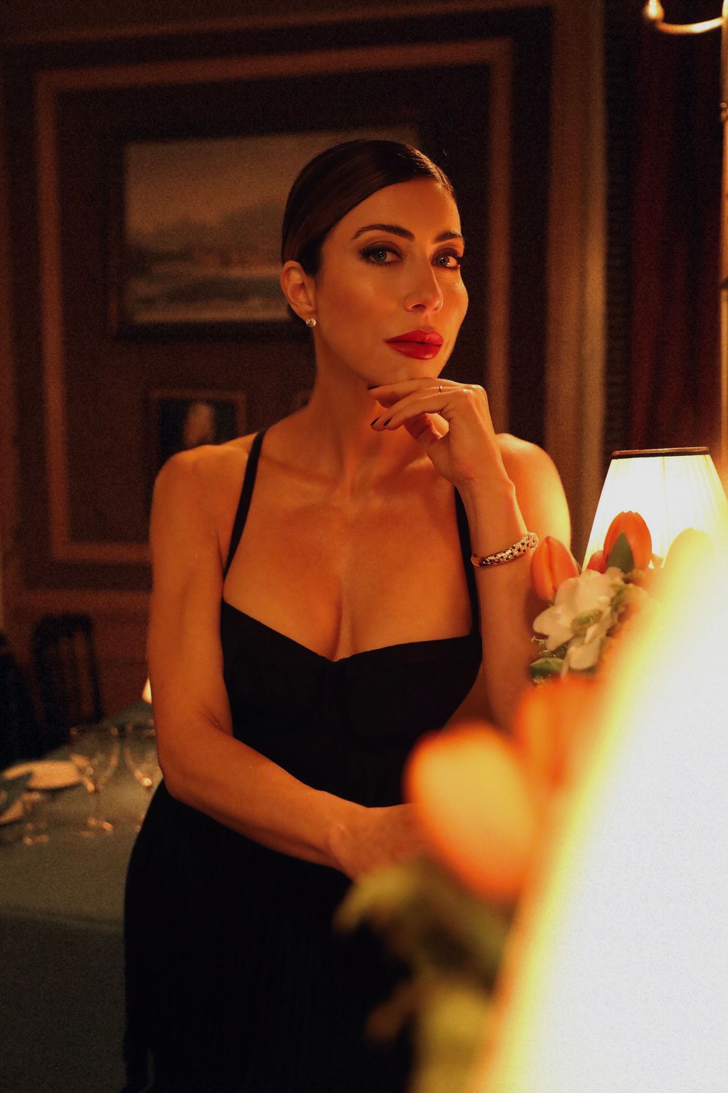 Moody portrait of a person in a black dress with red lipstick in warm candlelit setting.