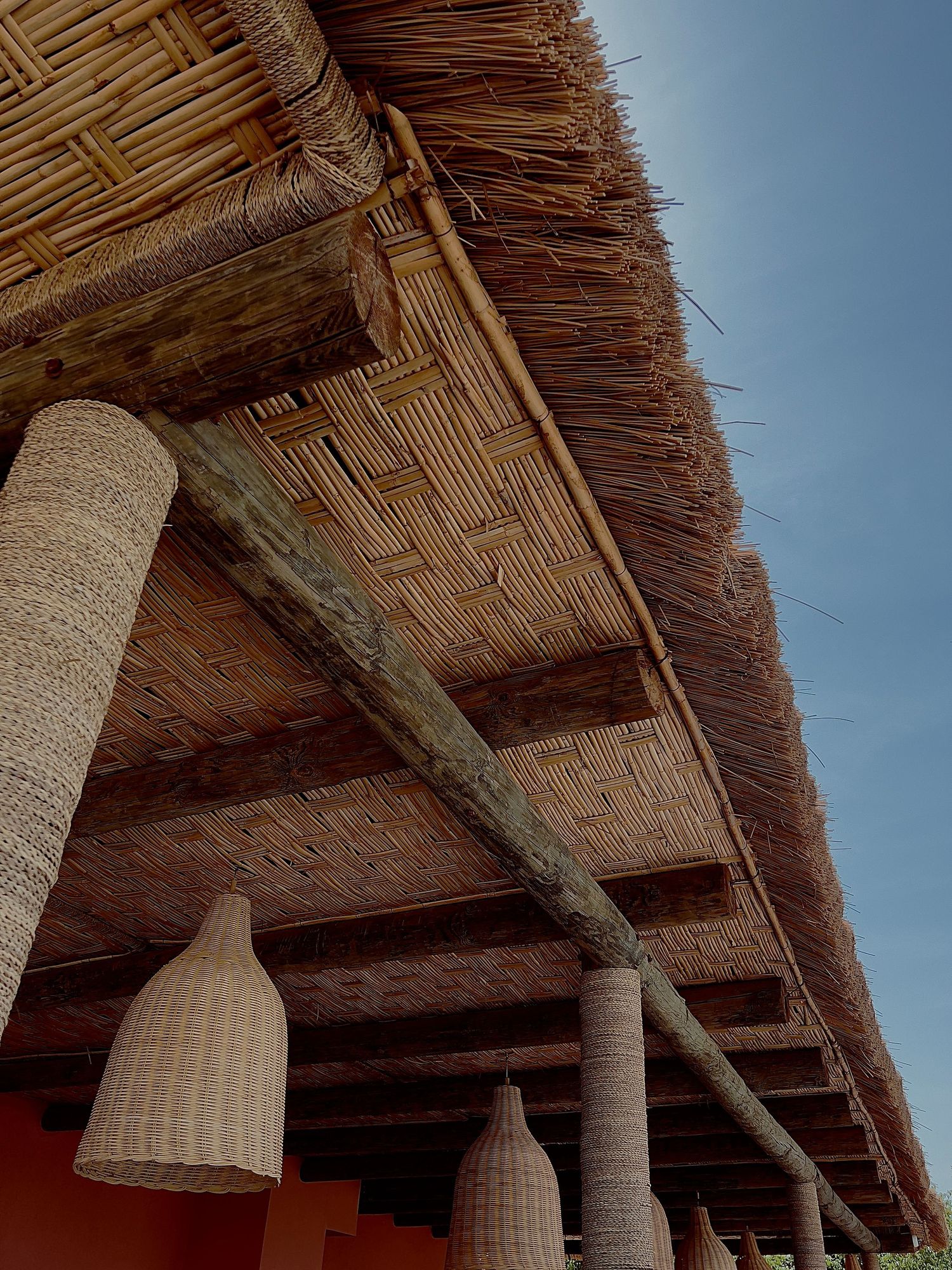 Hanging woven bamboo lamp shades under a thatched roof canopy against blue sky create a natural tropical aesthetic.