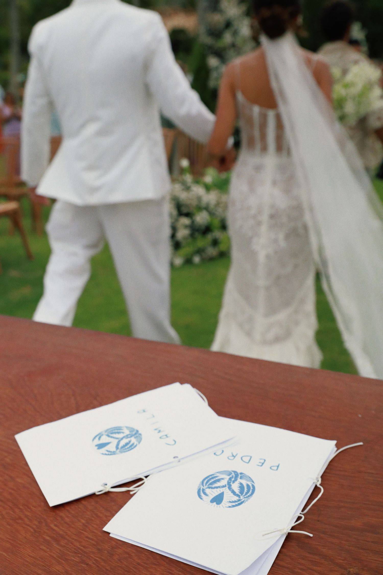Wedding ceremony programs with blue designs sit on wooden table as newlyweds walk in background on garden lawn.