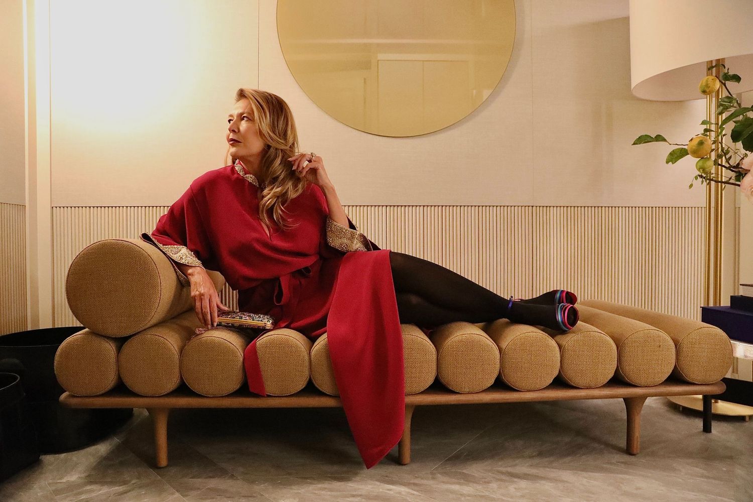 Someone in a red outfit reclines on a modern wooden daybed with cylindrical cushions in a minimalist room setting.