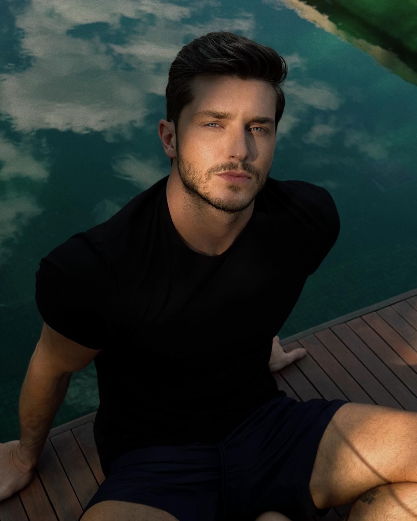 A person in black t-shirt poses by a swimming pool with reflective water during golden hour.