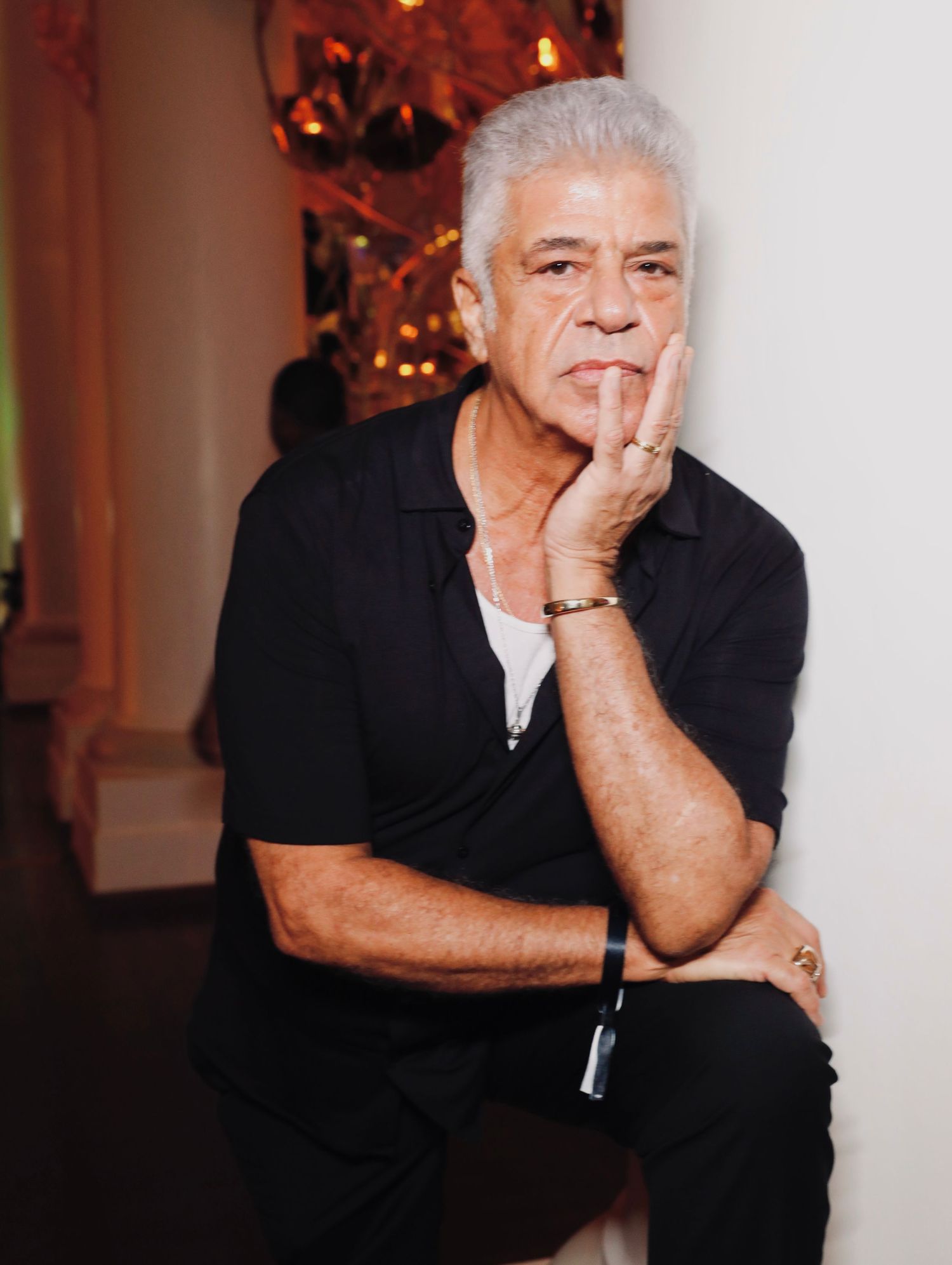 Mature man in black shirt seated thoughtfully in dimly lit room.