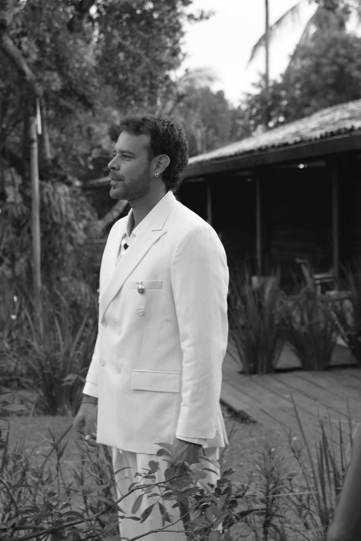 Black and white portrait of someone wearing a white suit jacket standing in a lush tropical garden setting.