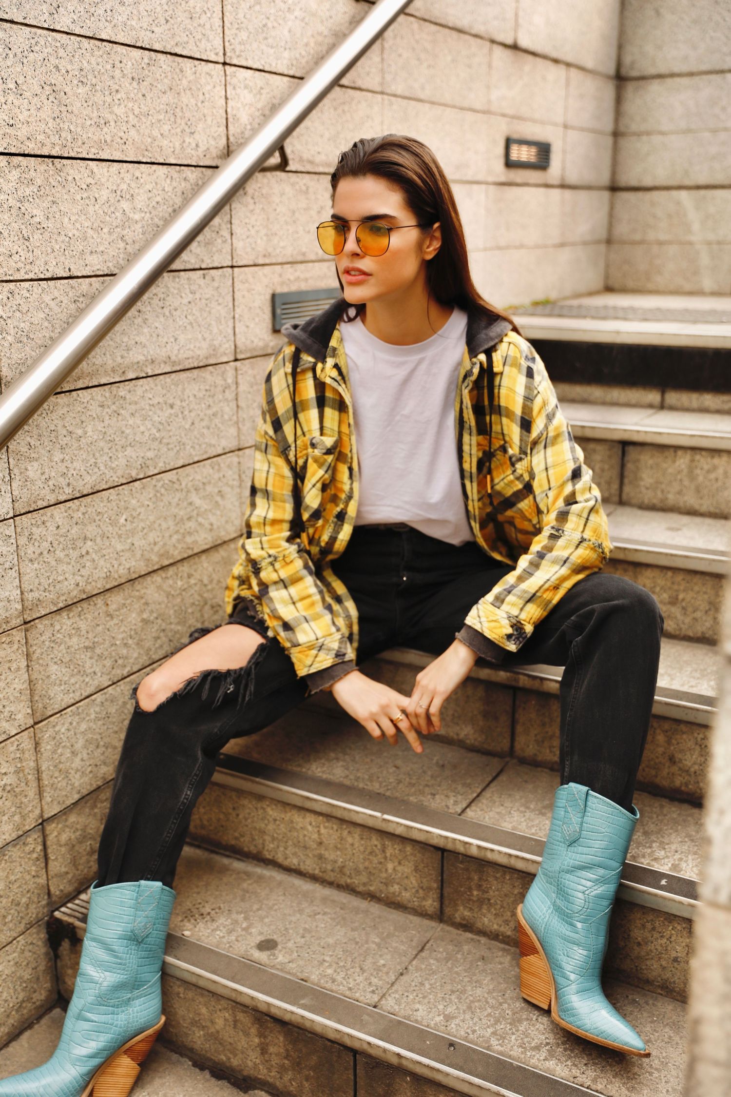 Person in trendy outfit with yellow plaid shirt, yellow sunglasses, white tee, and turquoise cowboy boots sits on concrete steps.