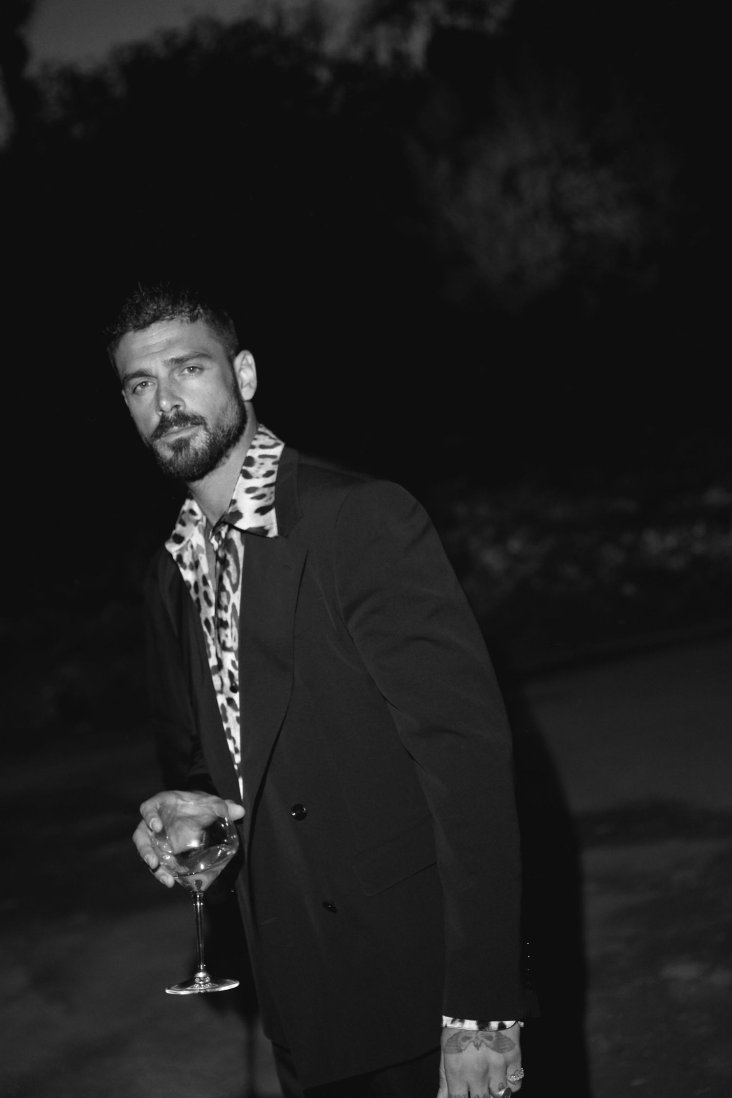 A black and white portrait of a person in formal attire holding a cocktail glass at night.