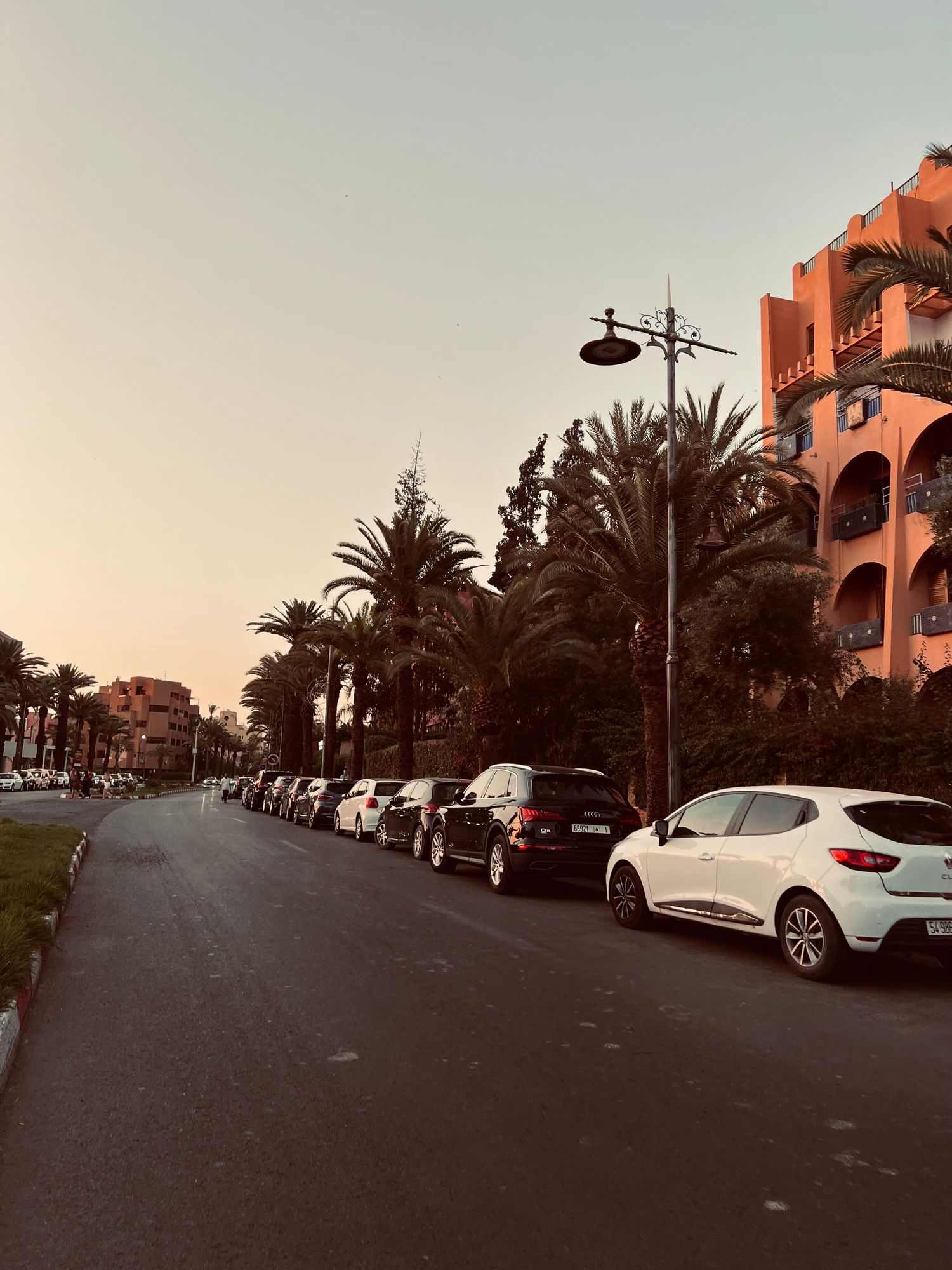 Street lined with palm trees and parked cars at sunset with Spanish-style buildings in a Mediterranean resort setting.