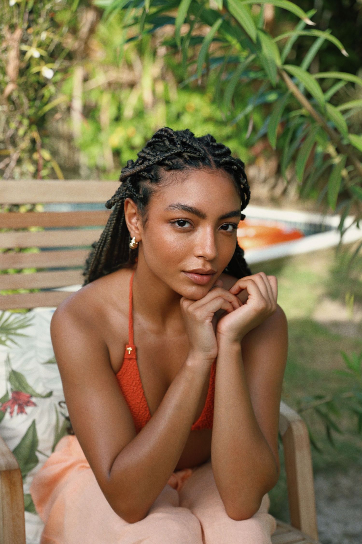 Portrait of someone with braided hair wearing orange swimwear against green tropical backdrop.