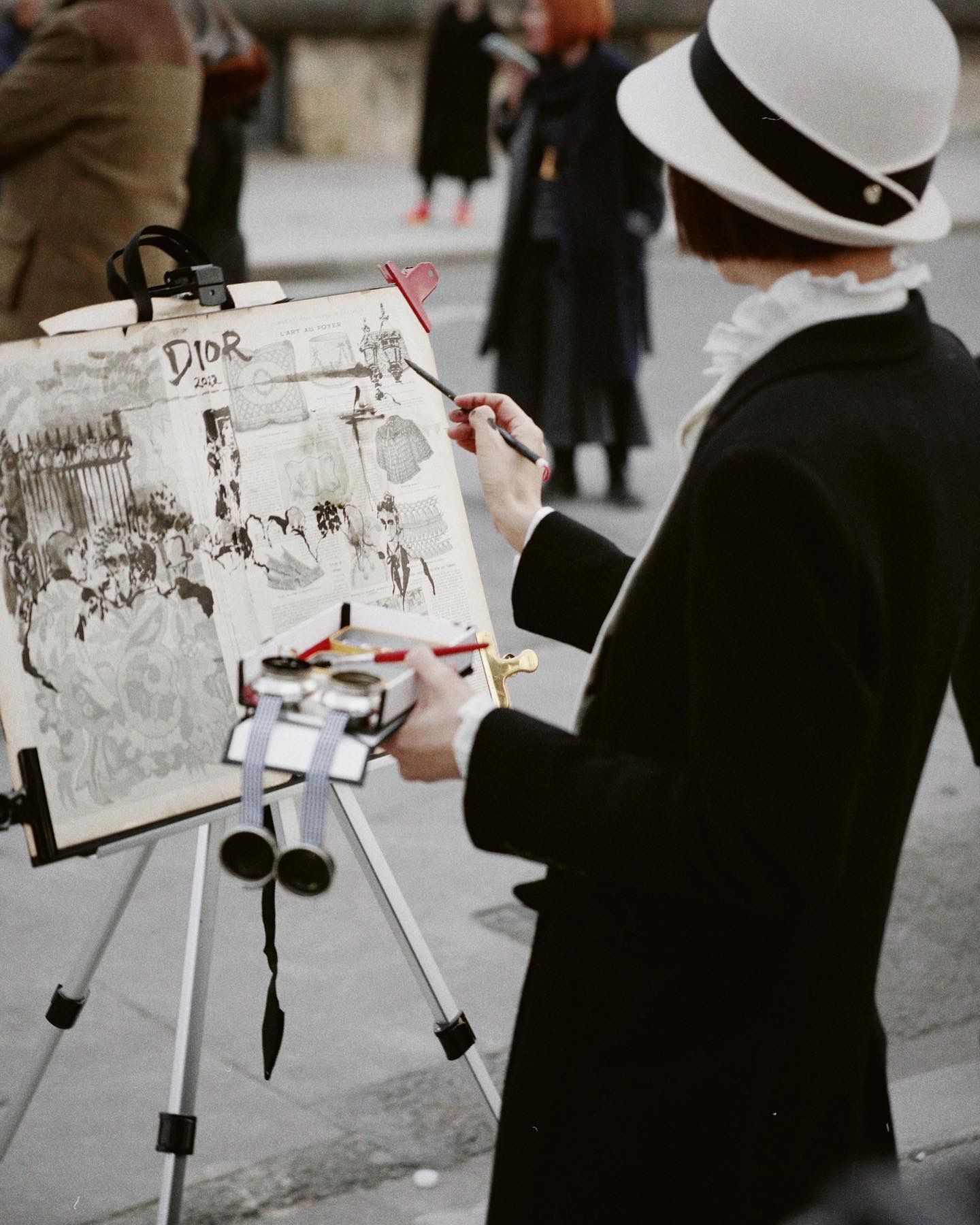 Artist in a white hat sketches on a white sketchbook mounted on a tripod outdoors in a public space.
