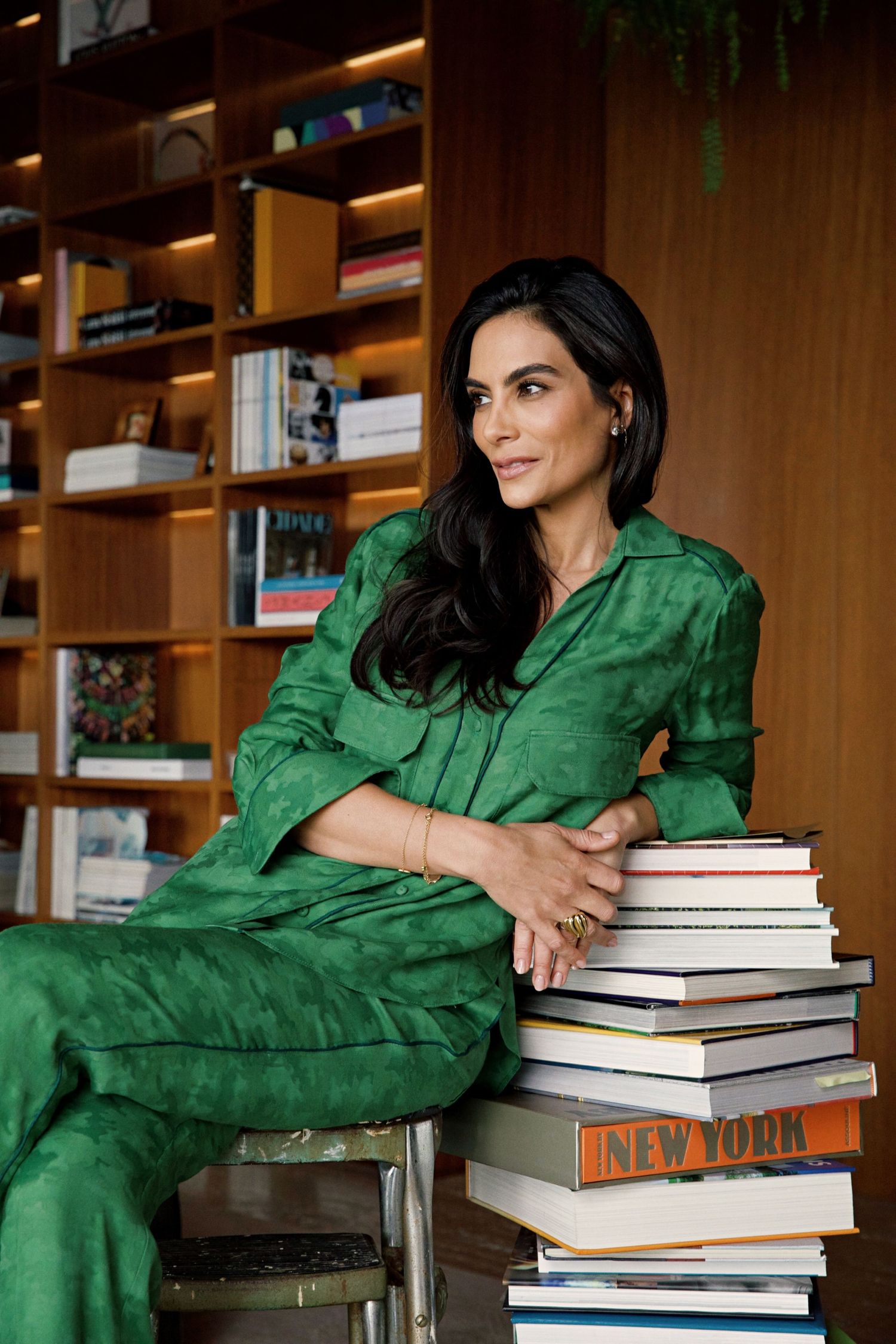 Person in emerald green outfit sits beside stack of books in modern home library setting.