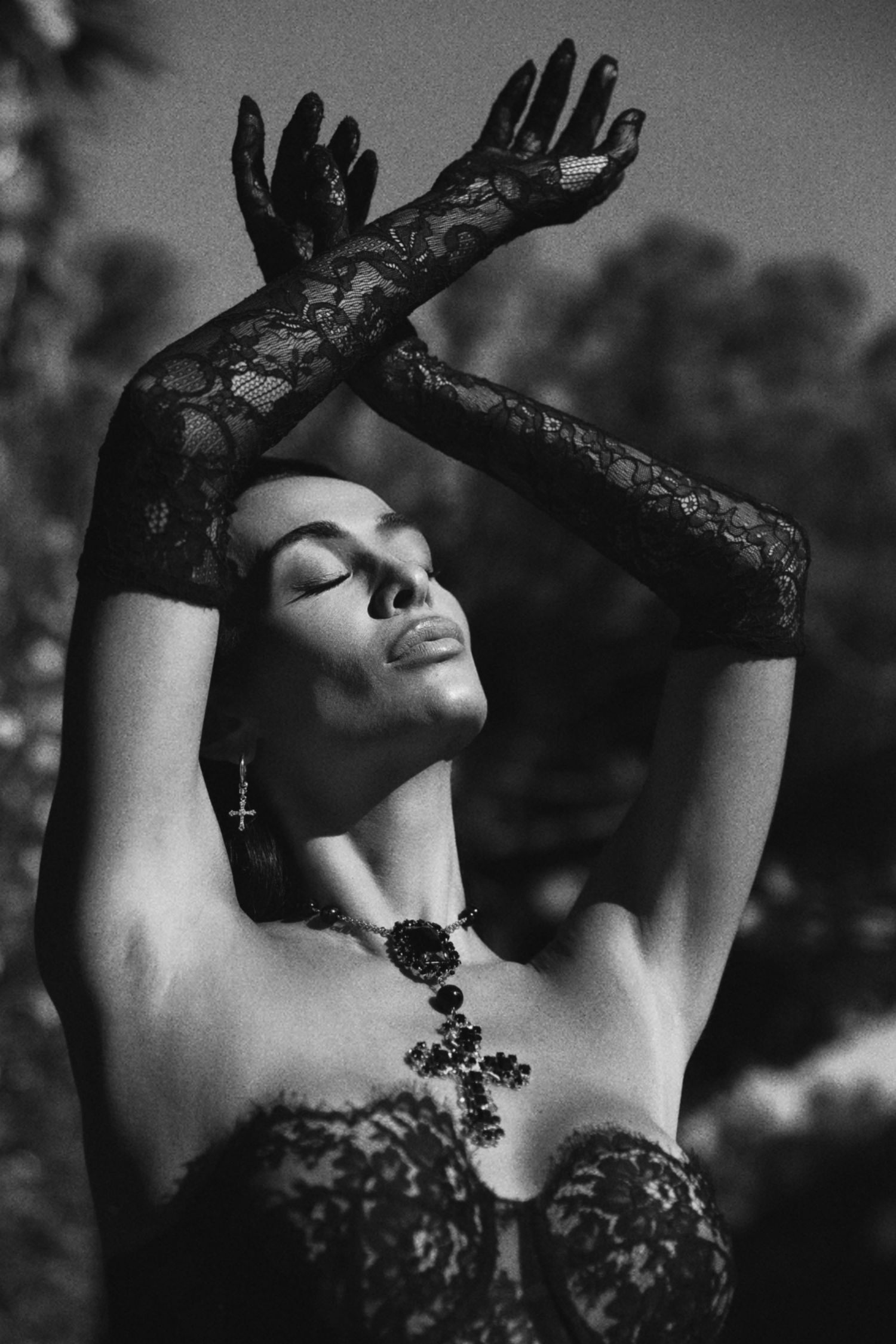 A dramatic black and white portrait of a figure in lace gloves and corset striking an elegant pose with raised arms.