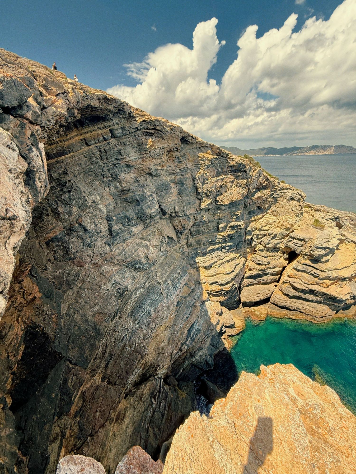 Dramatic rocky coastal cliffs tower above turquoise waters with white clouds billowing in the blue sky.