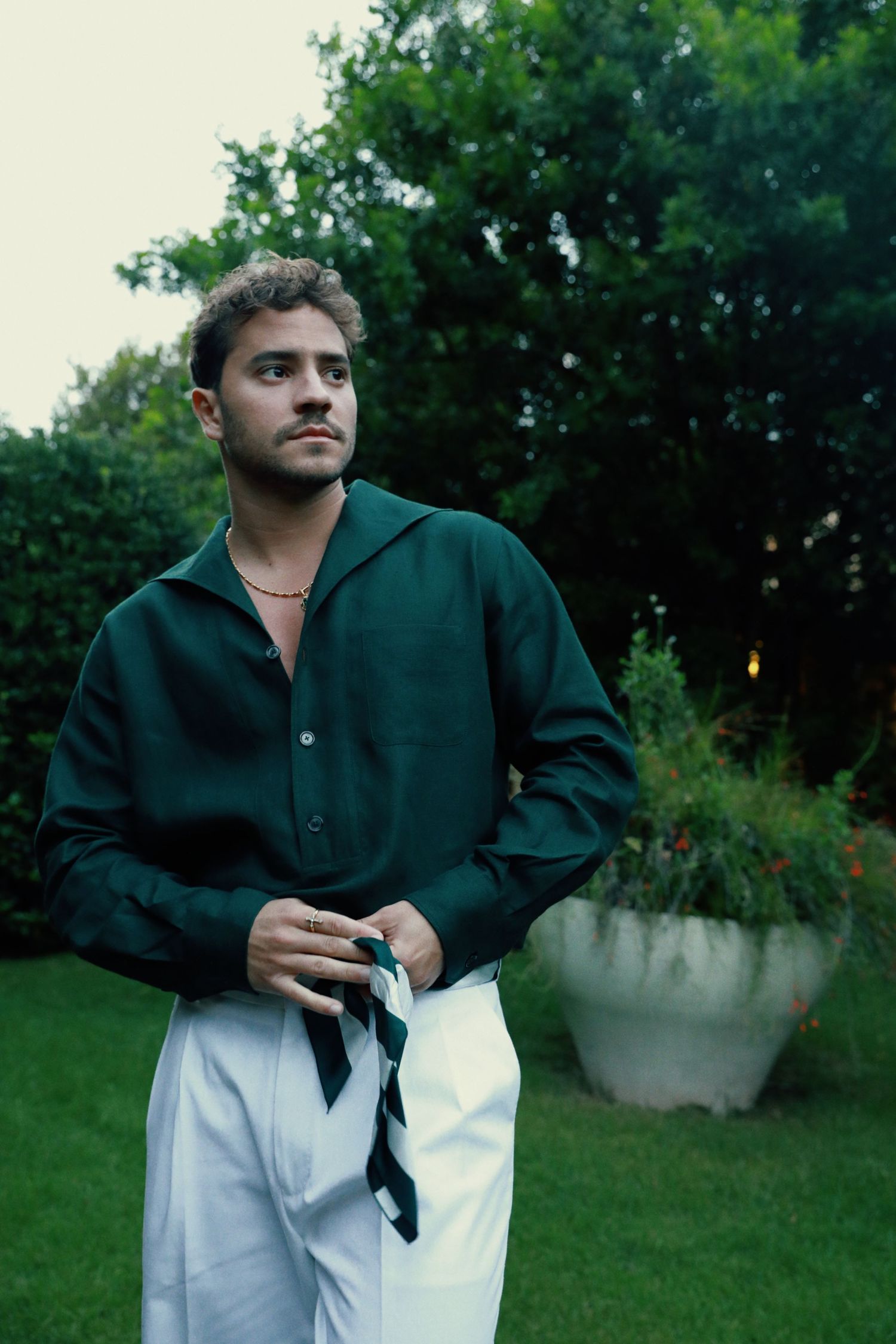 A person in a dark green button-down shirt and white pants stands in a garden setting during golden hour.