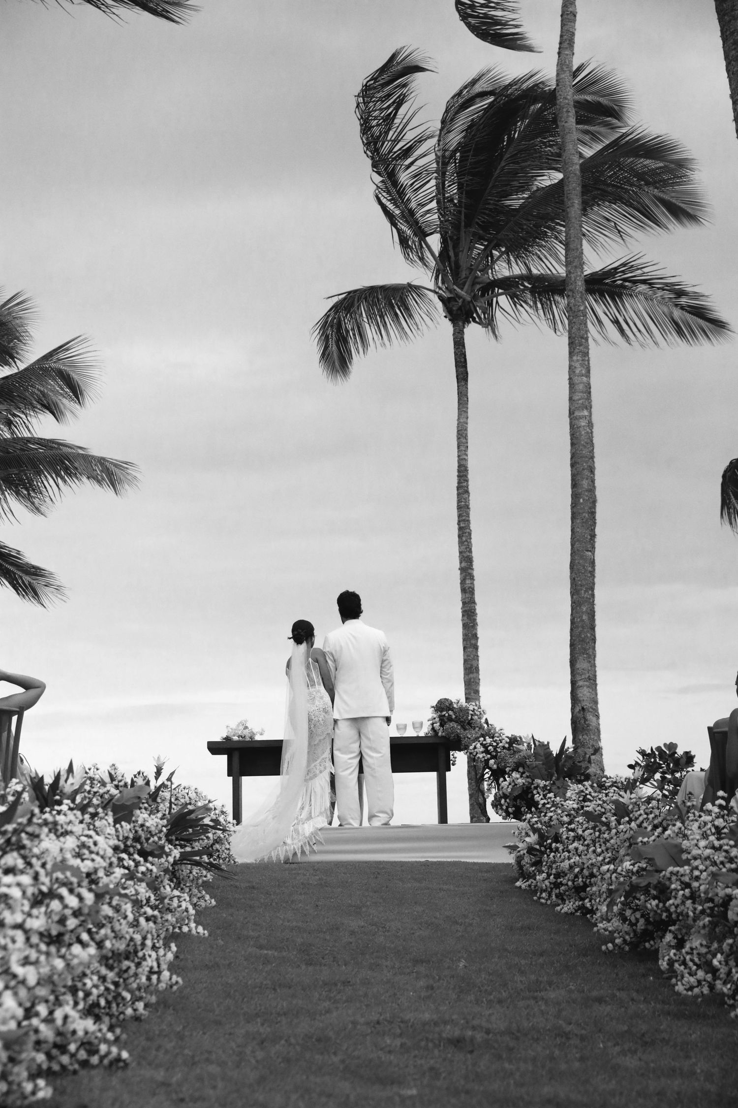 A couple in white stands on a tropical pathway lined with palm trees and flowers in black and white photography.