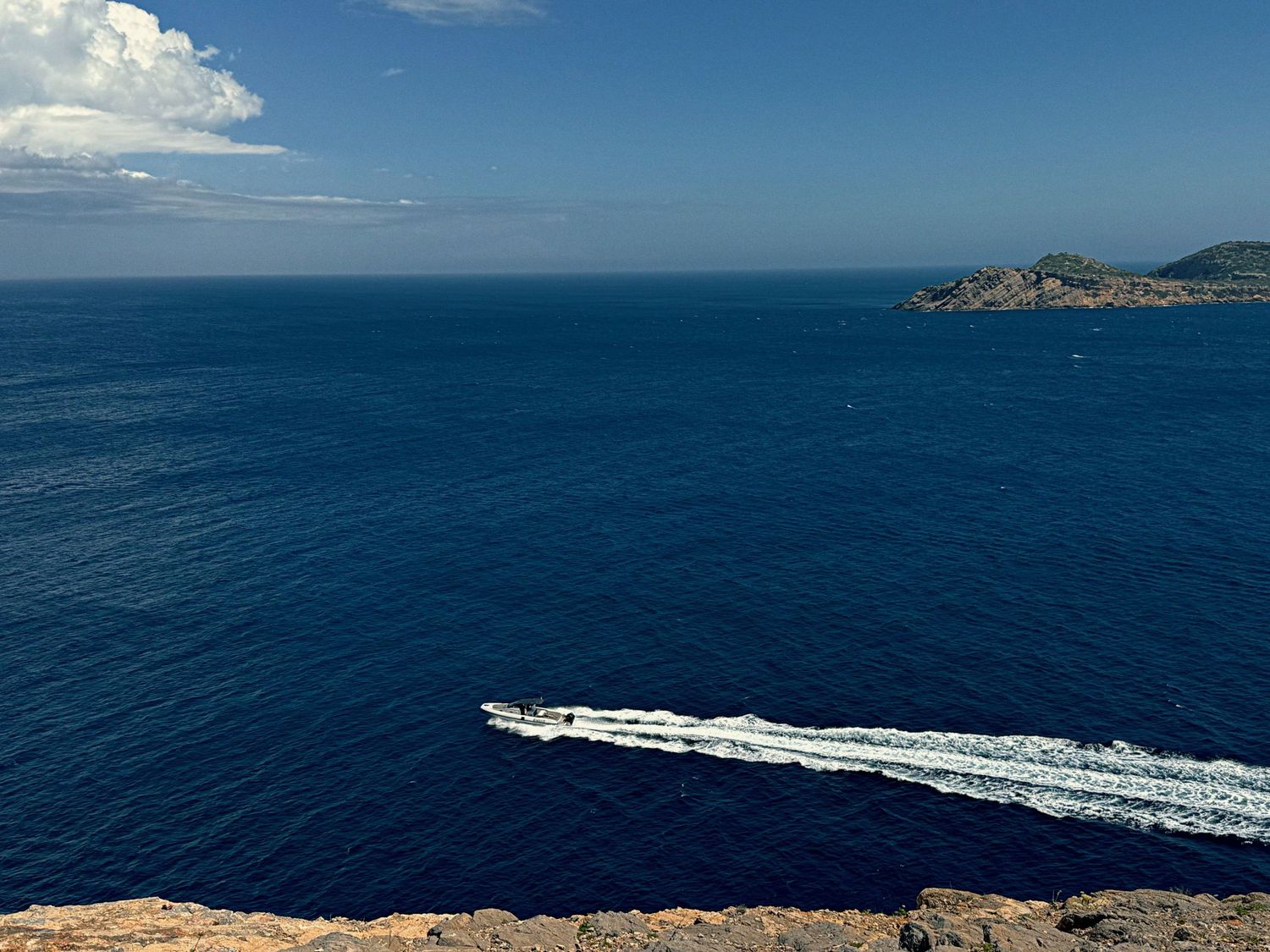 A speedboat creates a wake pattern in the deep blue Mediterranean waters near a rocky coastline.