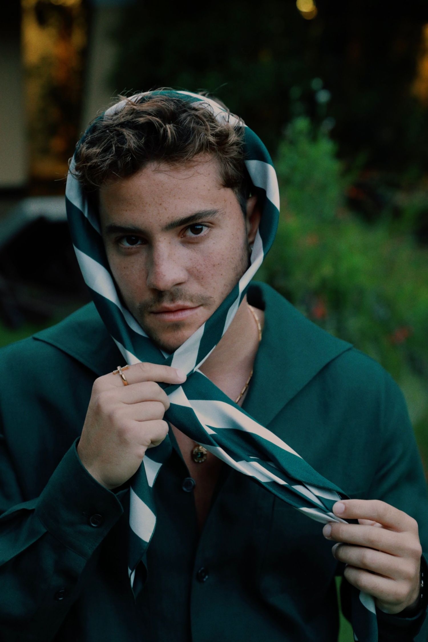 Person wearing a dark hoodie holds up a striped diagonal patterned scarf or tie against foliage background.