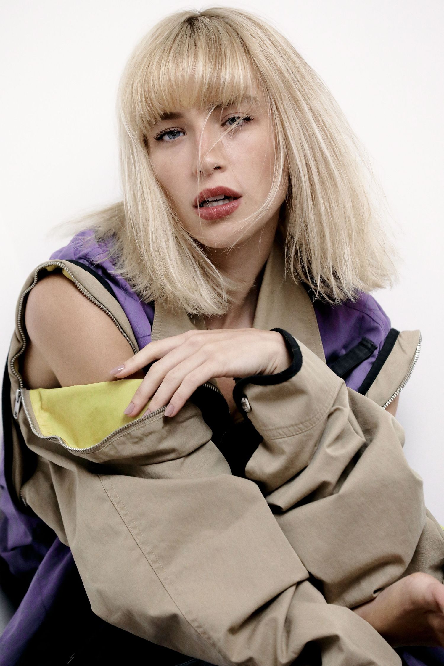 Fashion model wearing a beige jacket with purple accents and blonde bob hairstyle poses against white background.