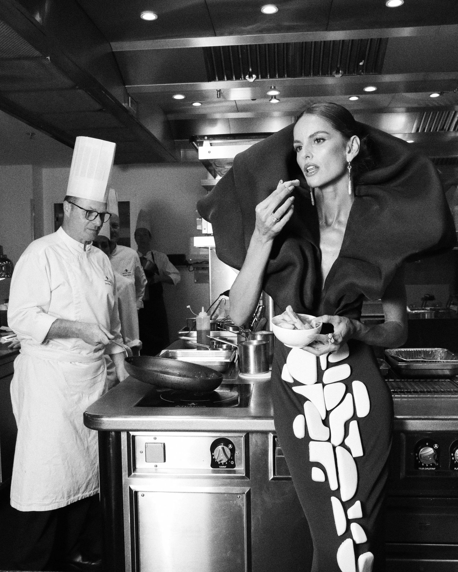 Black and white photo of model tasting food in geometric dress with chef working in background.