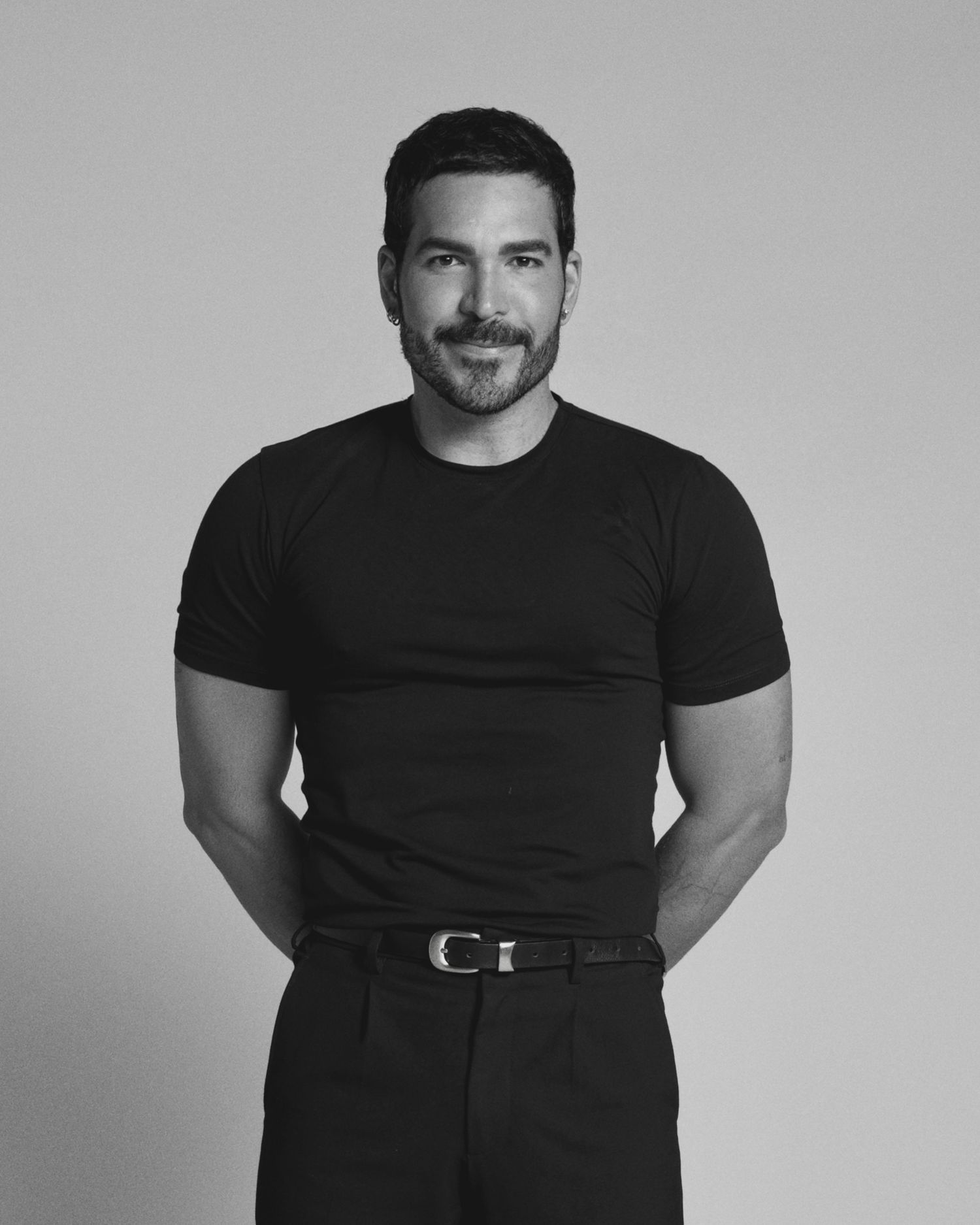 Black and white studio portrait of a man in fitted black t-shirt and pants with belt.