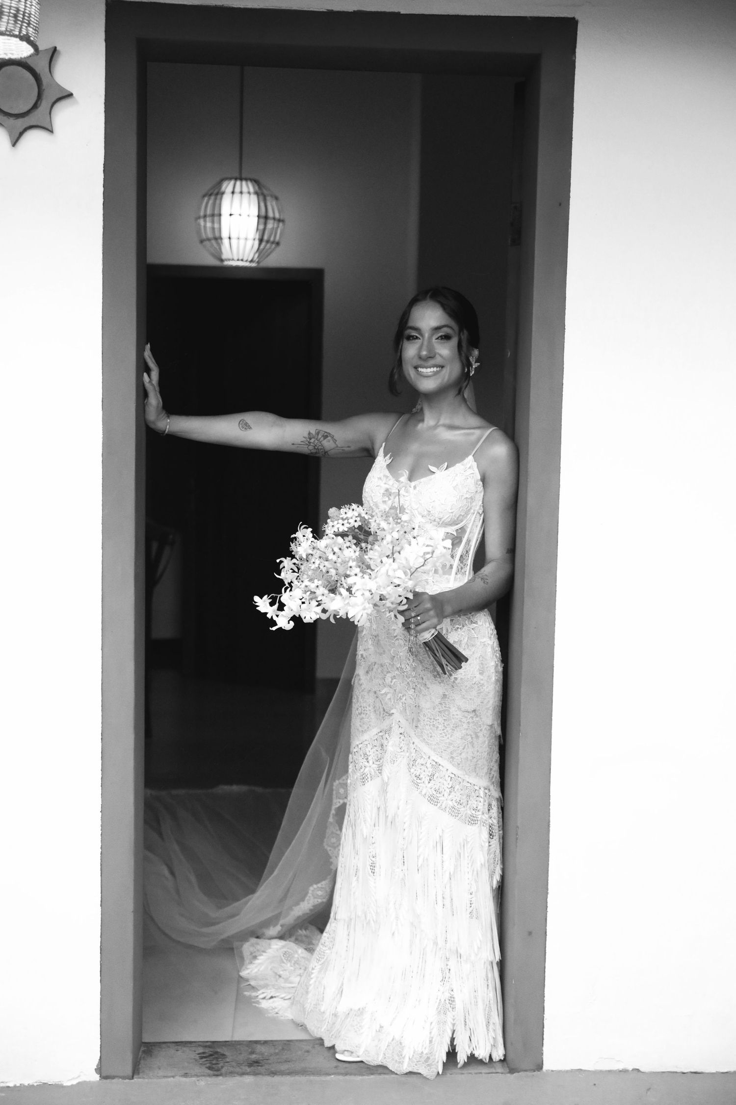 A bride in a white lace wedding dress with spaghetti straps poses in a doorway holding a bouquet of white flowers.