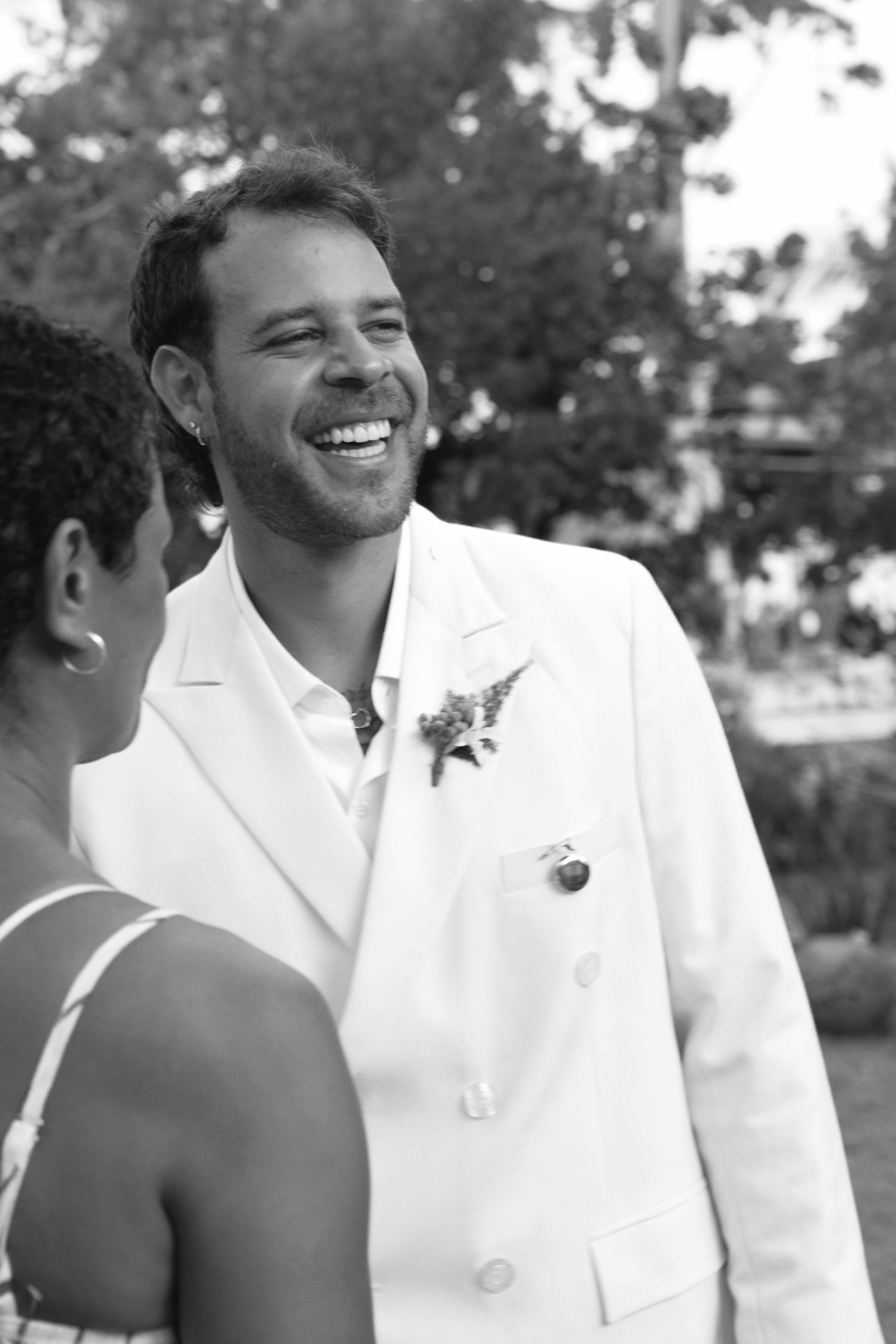 Black and white photo of someone in a white suit jacket with a boutonniere smiling during a joyful outdoor moment.
