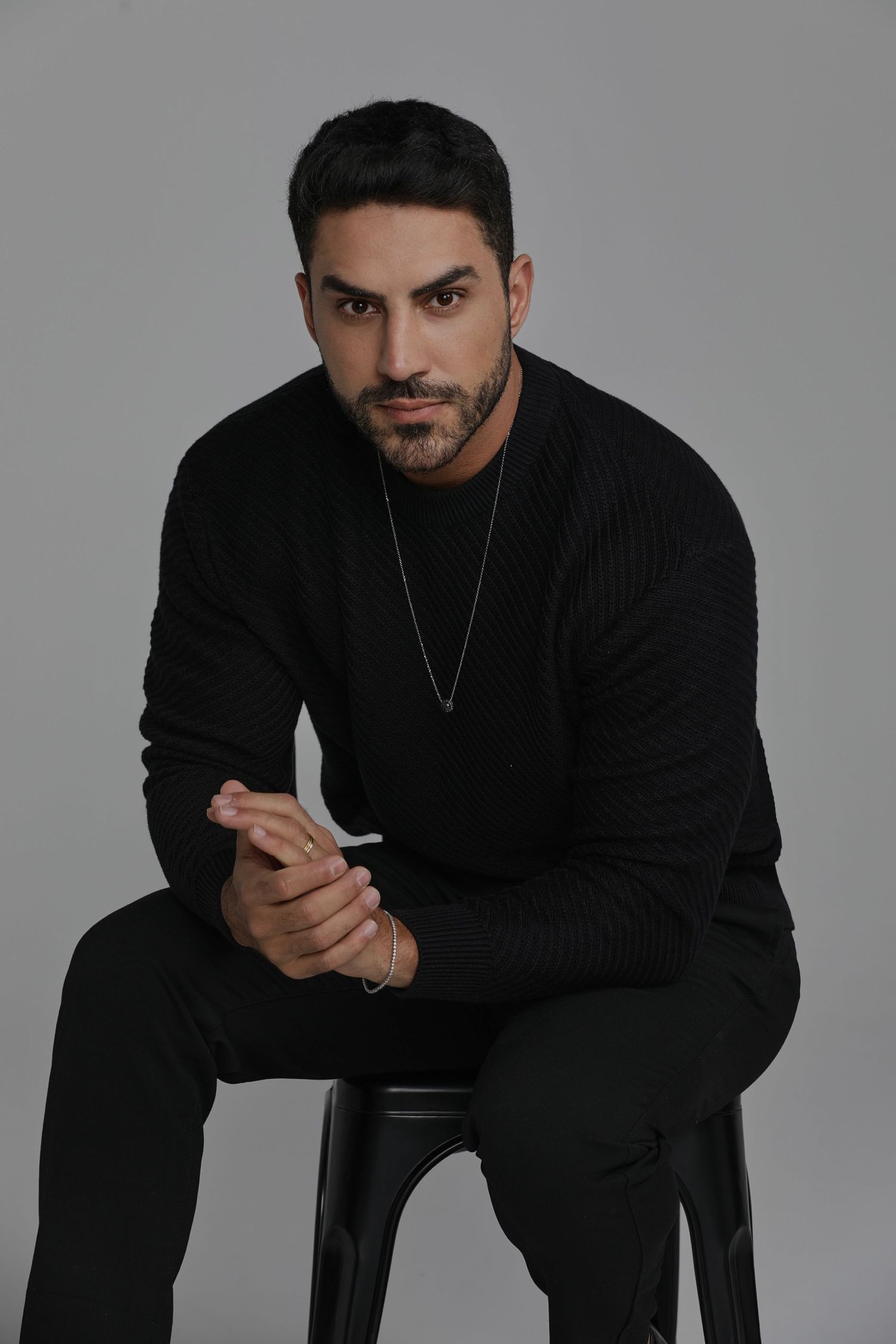 Portrait of man in black sweater seated on stool wearing silver necklace.