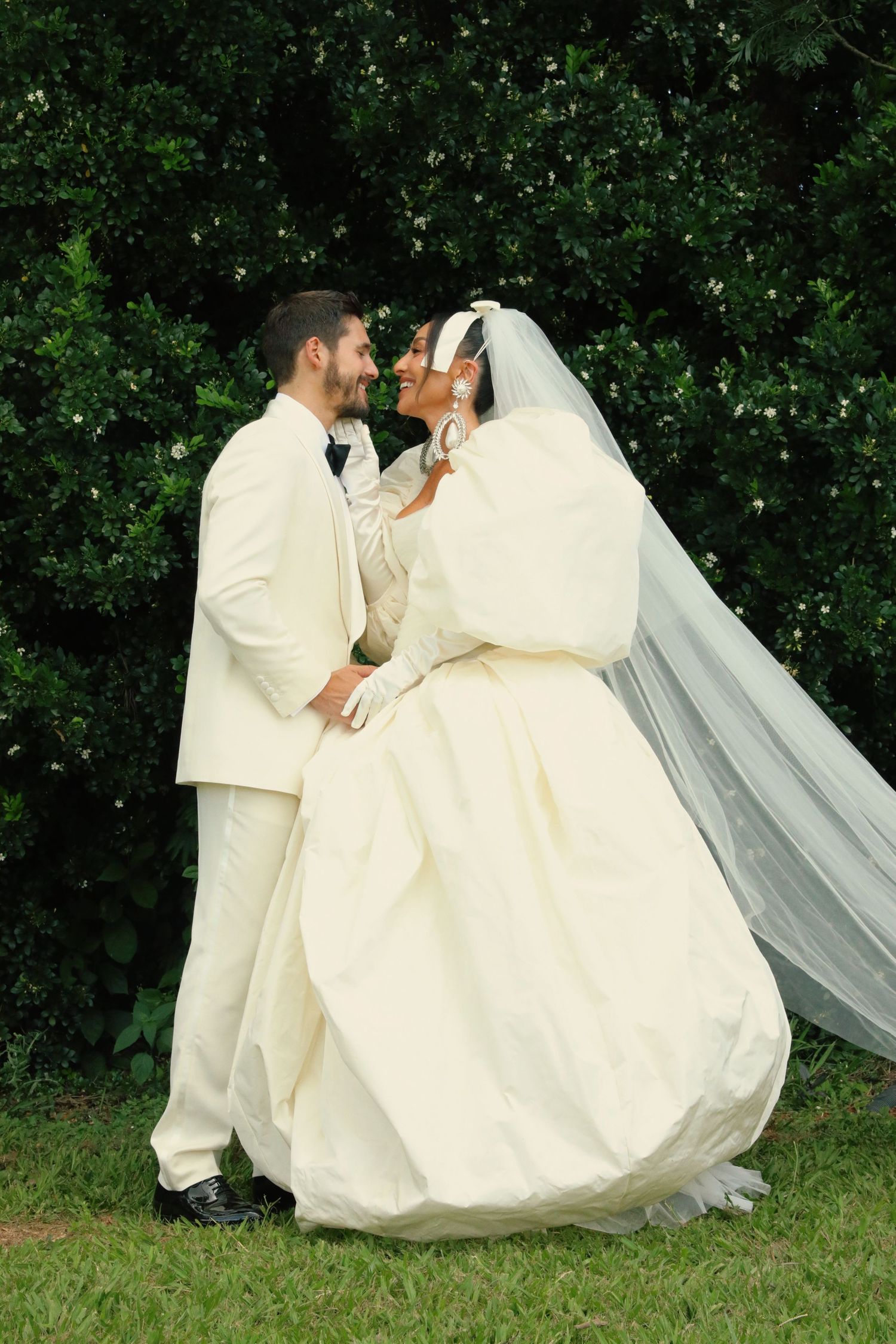 A tender moment between newlyweds in white attire against a backdrop of green leaves.