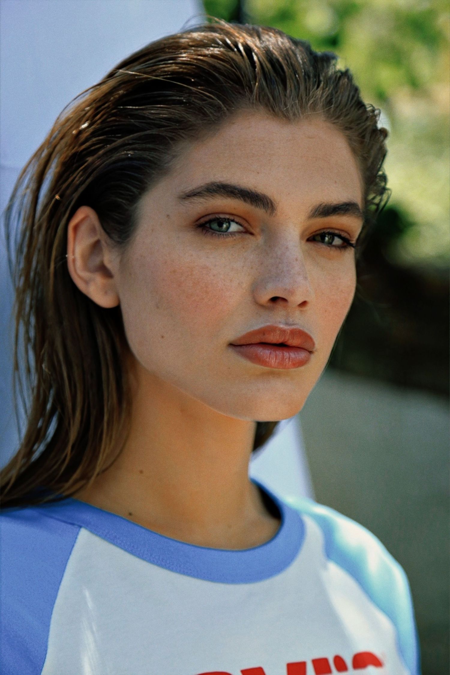 Portrait of a model with slicked-back dark hair wearing a blue and white t-shirt against a blurred outdoor background.