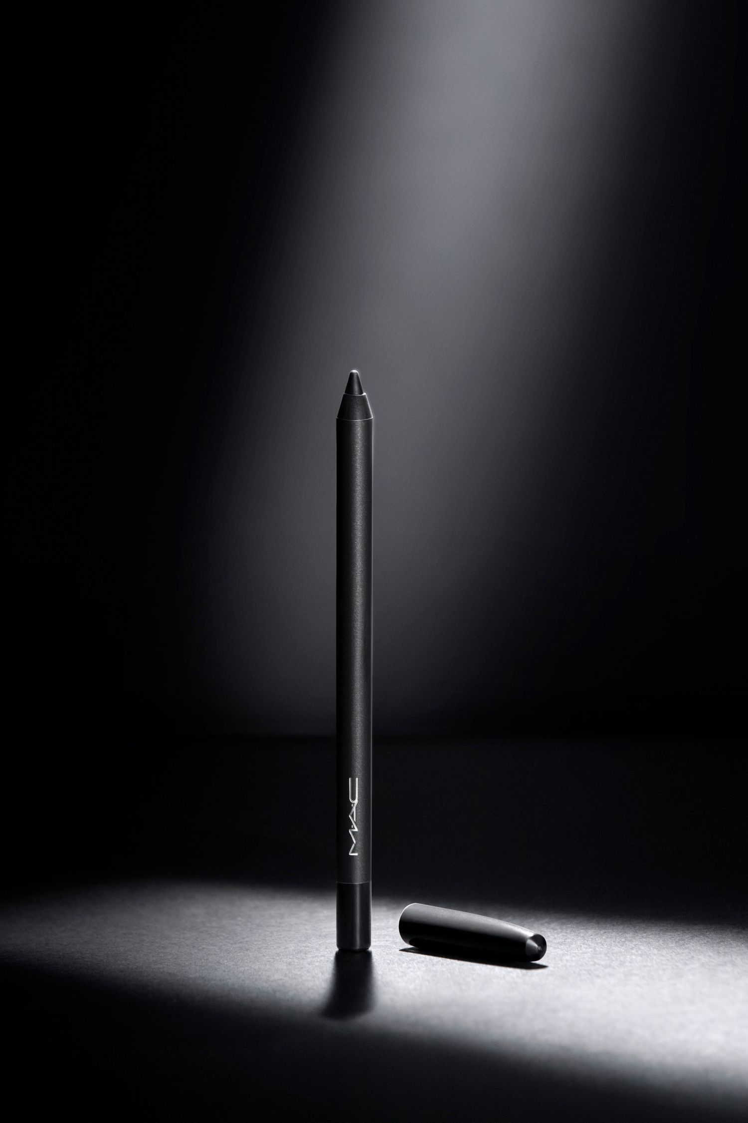 Black makeup pencil and cap photographed dramatically on dark surface with spotlight.